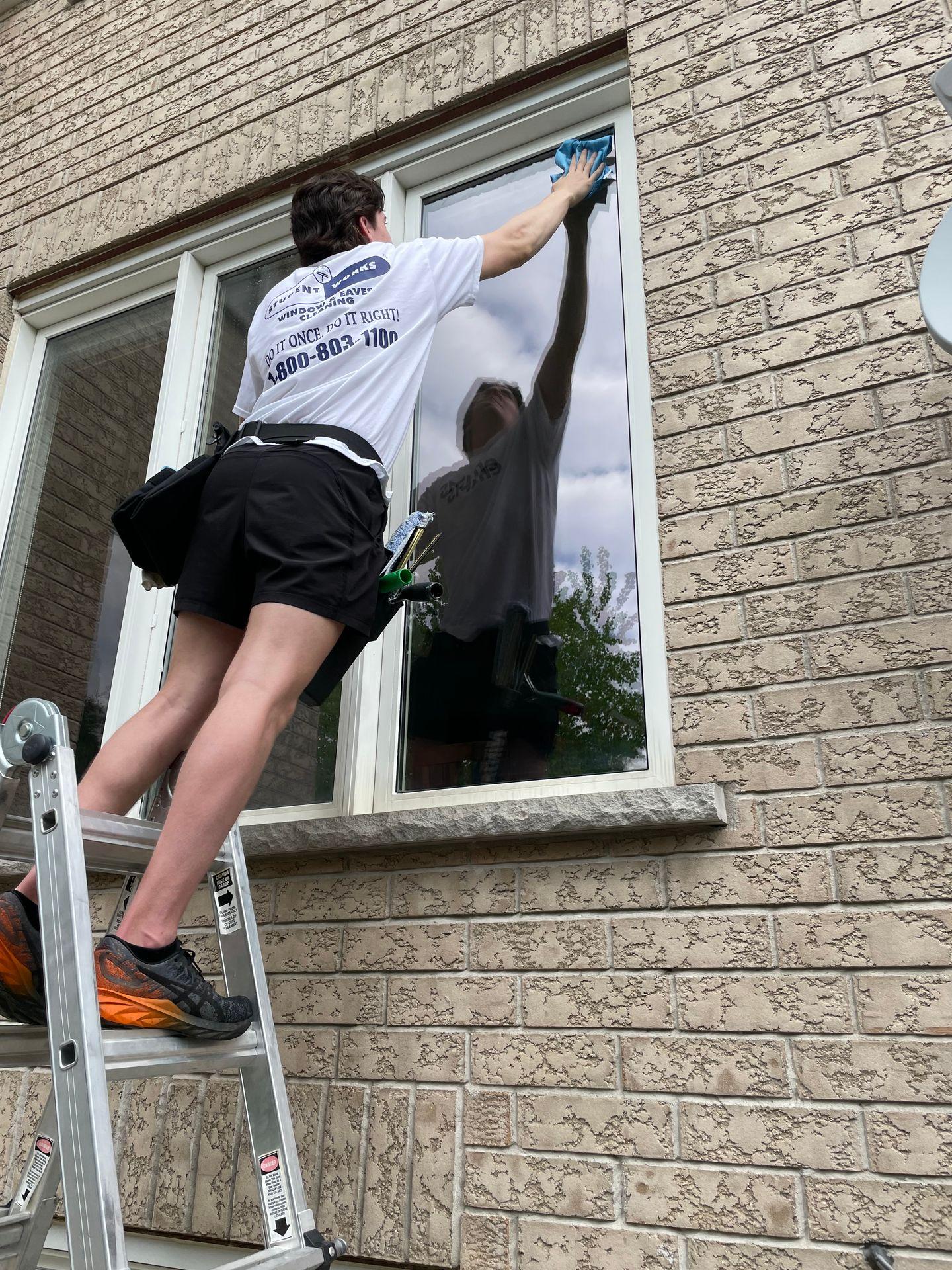 Person on a ladder cleaning a window with a cloth next to a brick wall.