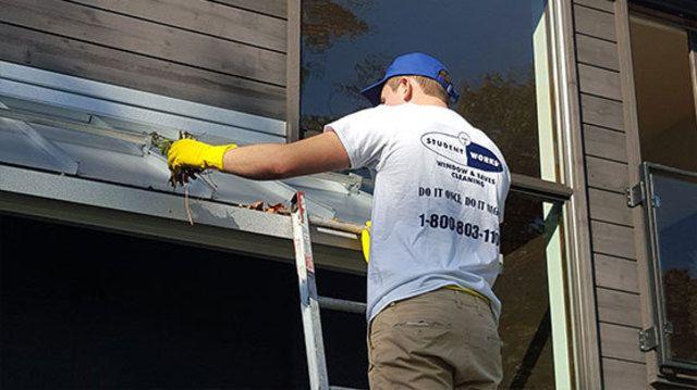 Person on ladder cleaning house gutters wearing gloves and a white work shirt.